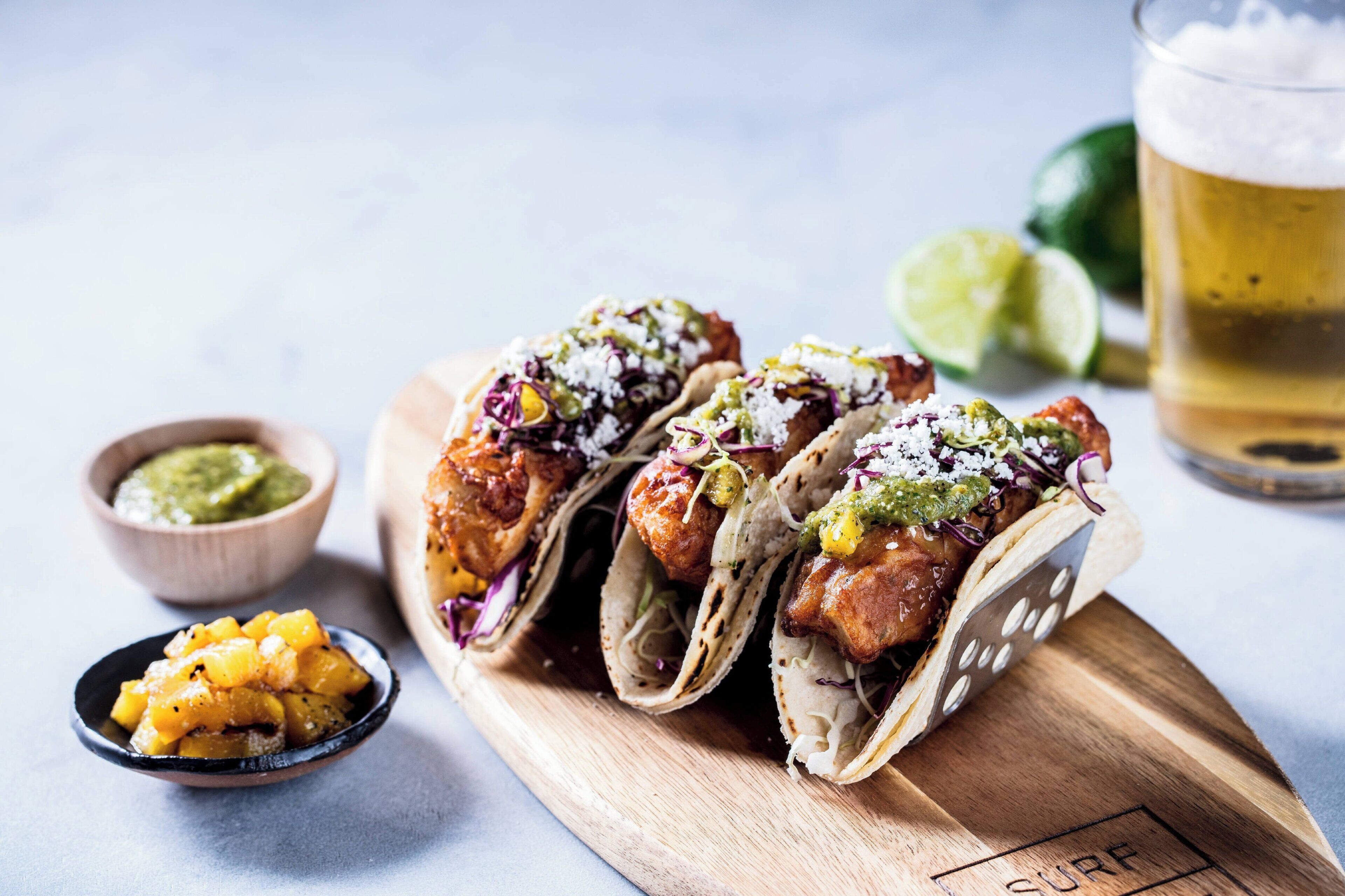 Three tacos on a wooden board with a glass of beer and lime wedges in the background.