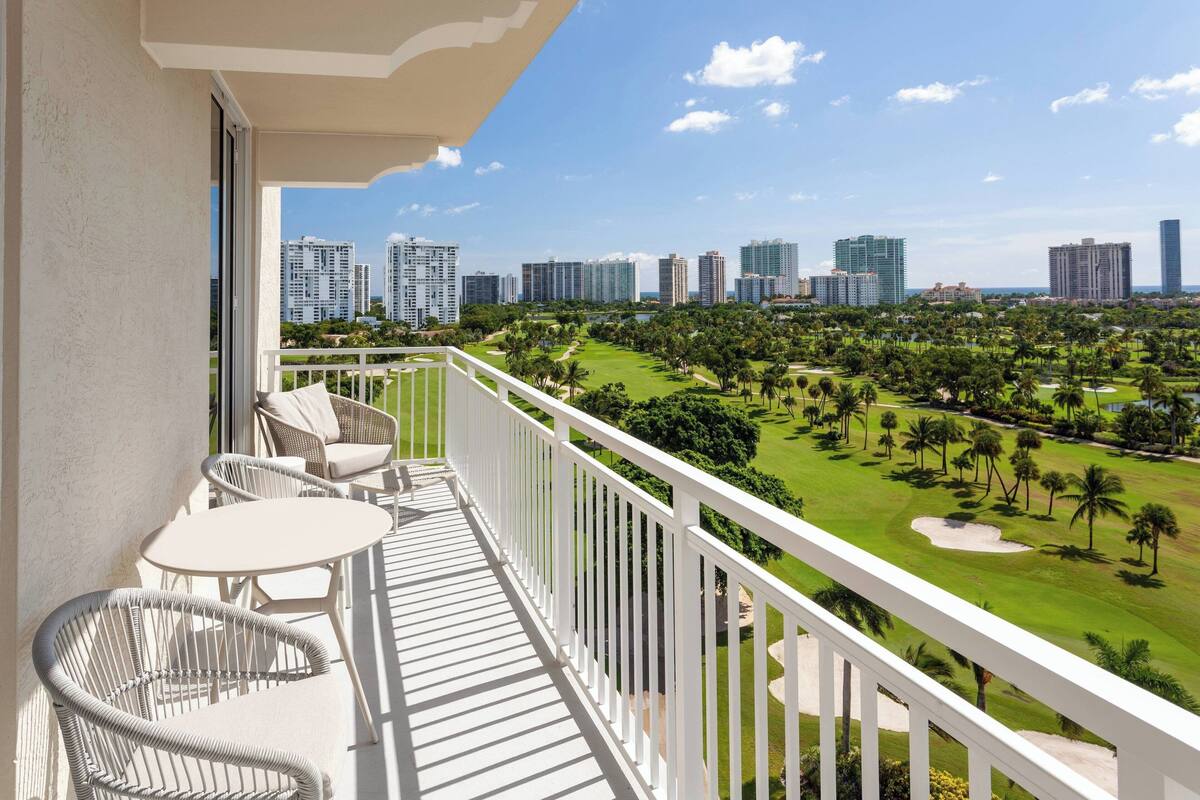 A balcony with white wicker chairs and a table, overlooking a golf course and city skyline.