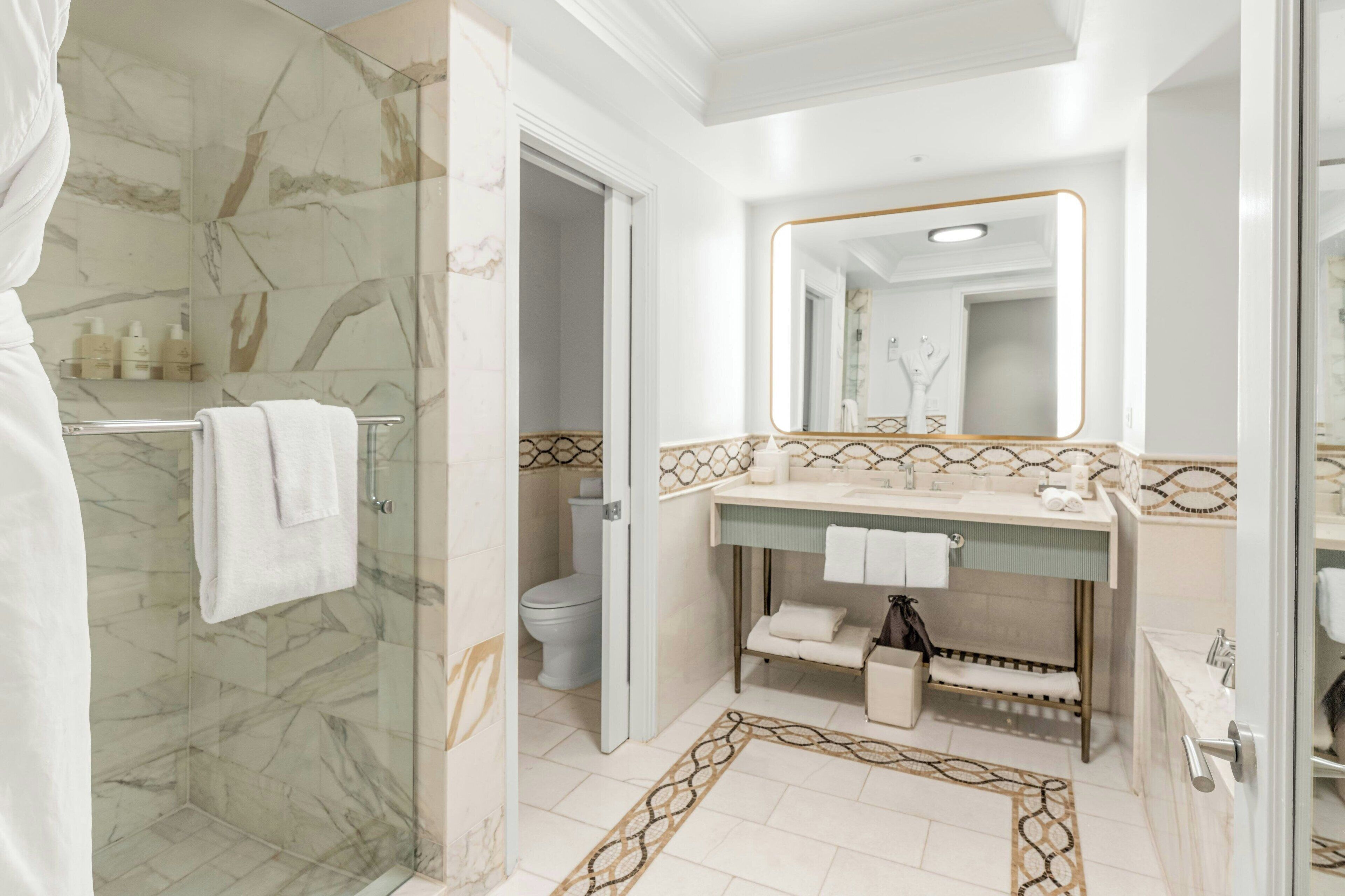 A bathroom with a glass shower enclosure, a marble-tiled floor, a vanity with a mirror, and a toilet.