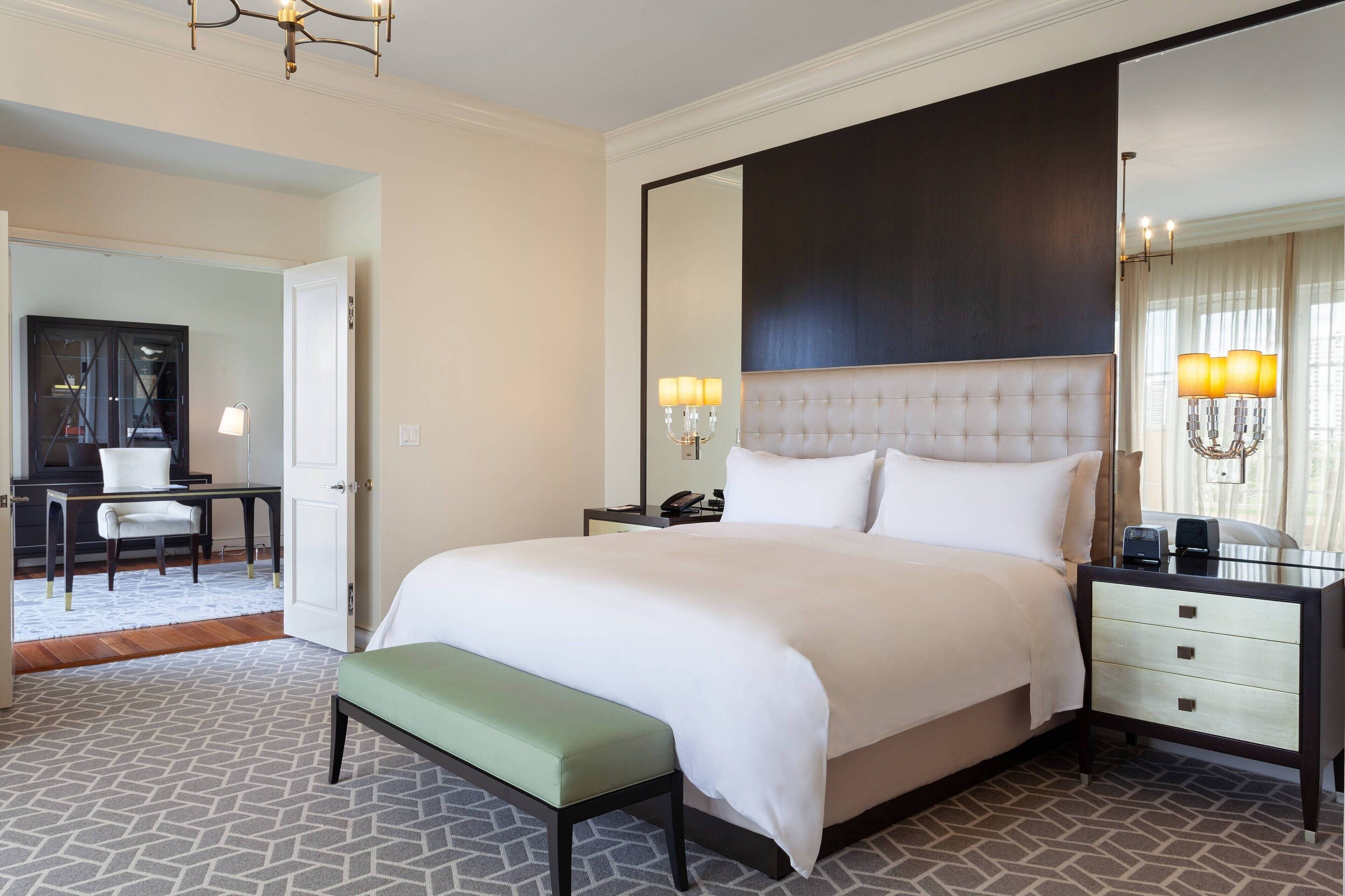A hotel room with a large bed, a bedside table with a lamp, a mirror, and a bench.