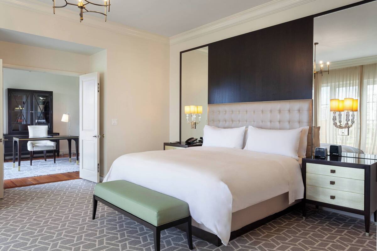 A hotel room with a large bed, a bedside table with a lamp, a mirror, and a bench.