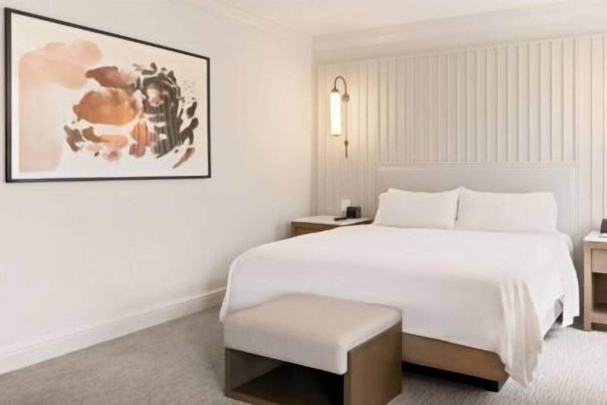 A hotel room with a large bed, bedside tables, a bench, a framed artwork, and a lamp.