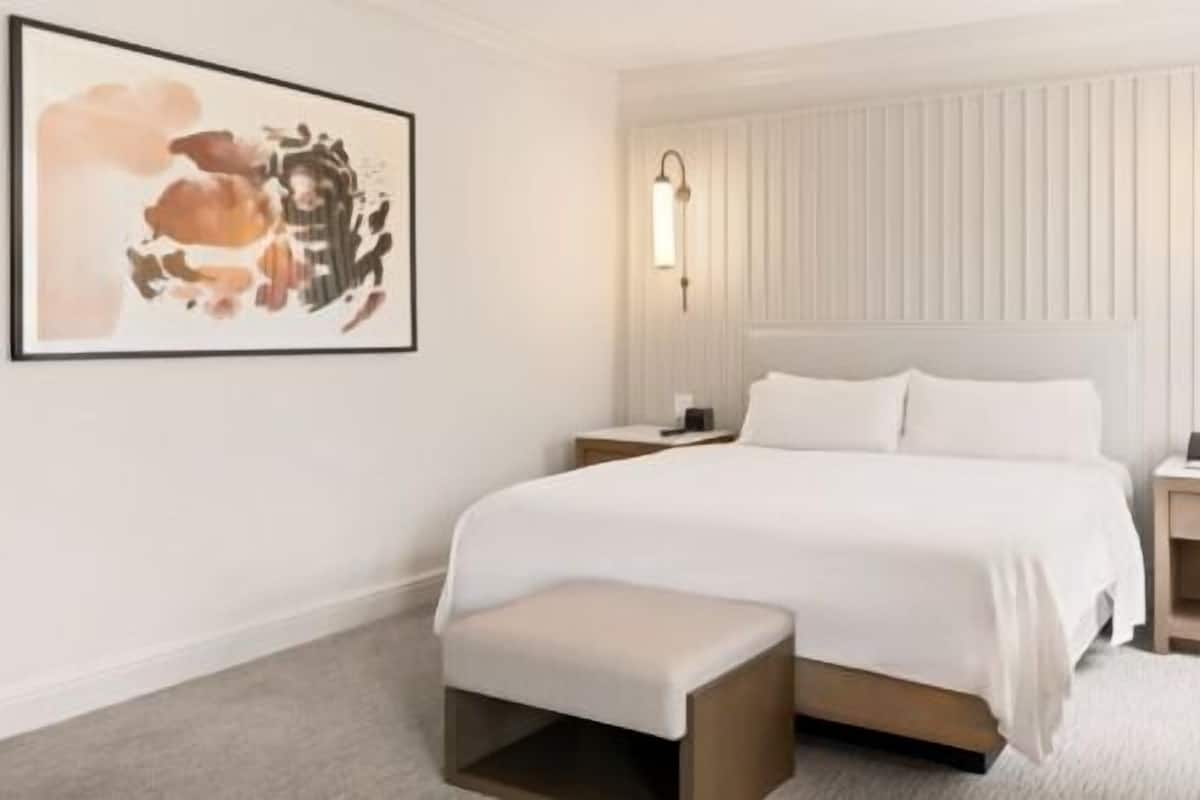 A hotel room with a large bed, bedside tables, a bench, a framed artwork, and a lamp.