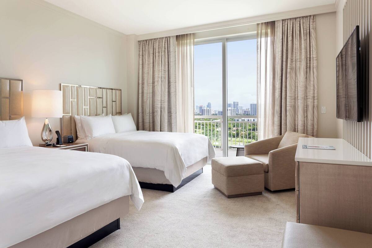 A hotel room with two beds, a desk, a chair, and a large window with a city view.