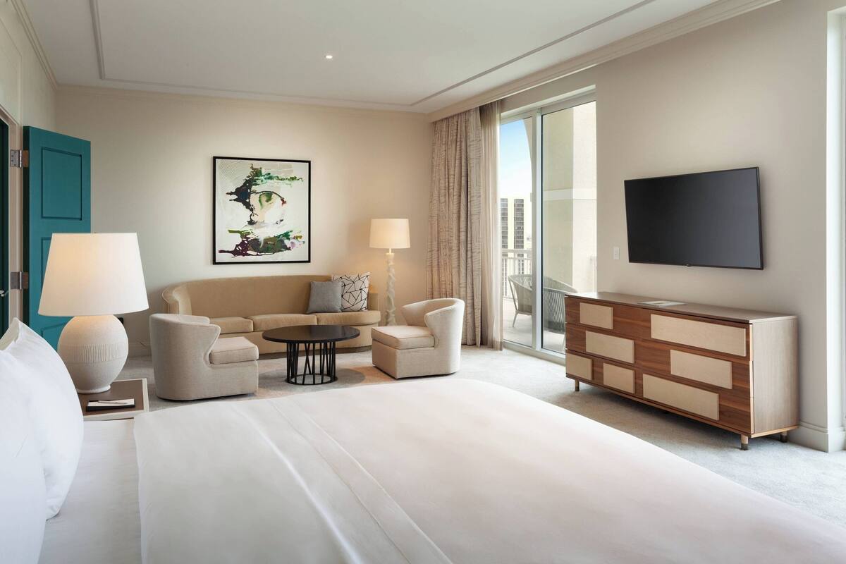 A hotel room with a large bed, a sofa, a coffee table, a TV, and a view of the city through the window.