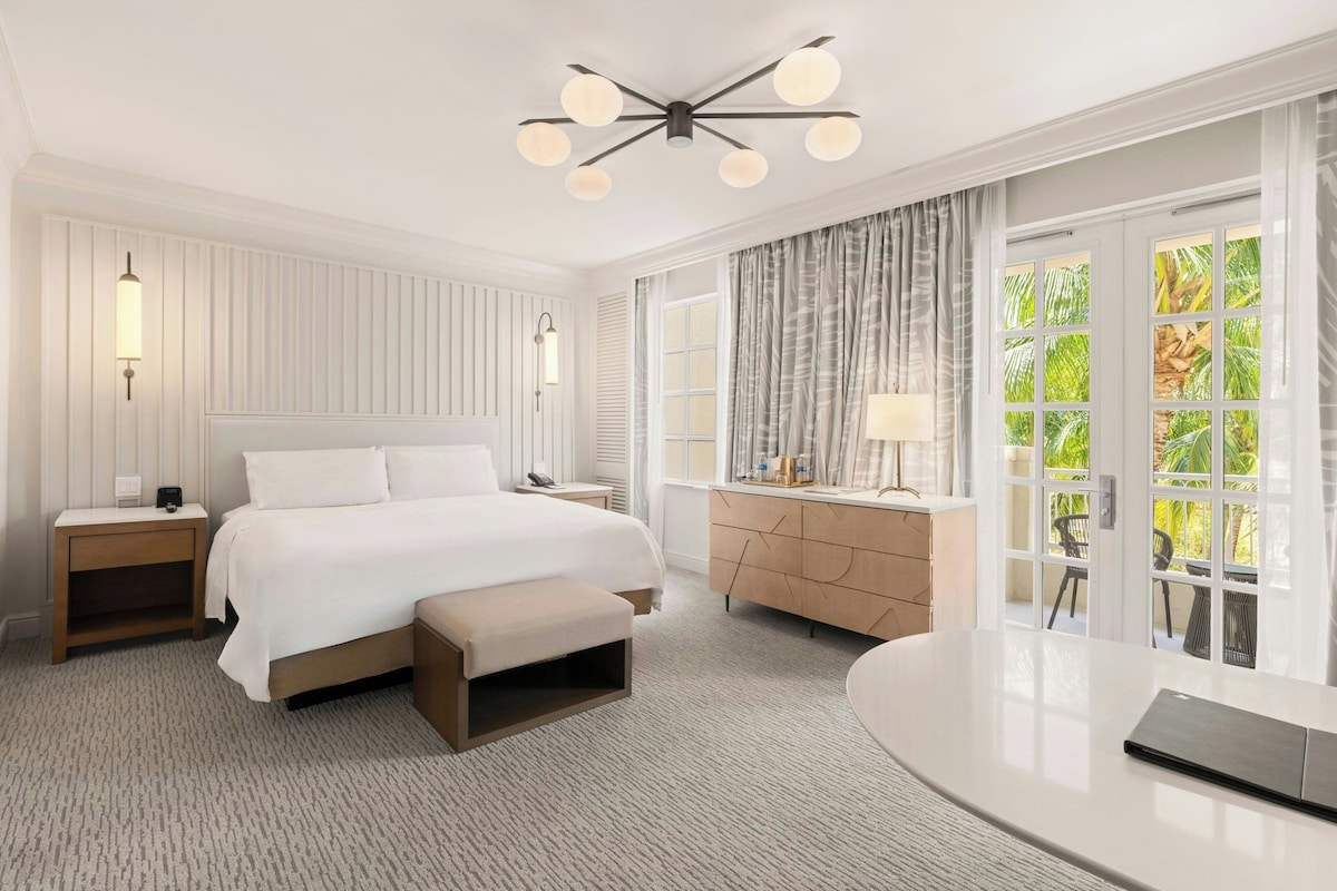 Ein modernes Hotelzimmer mit einem großen Bett, Nachttischen, einem Schreibtisch und Blick auf Grün durch große Fenster.