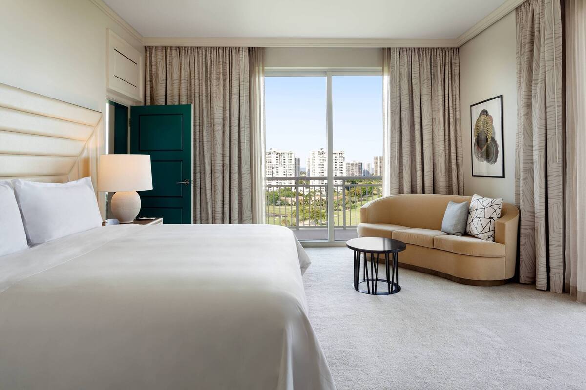 A hotel room with a large bed, a sofa, a small table, and a view of the city through large windows.