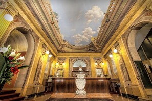 An ornate lobby with a grand chandelier, a large wooden desk, and classical architectural details.