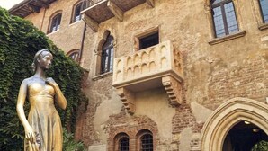 Un edificio con una estatua de una mujer vestida, ventanas arqueadas y un balcón con barandillas de piedra.