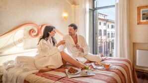Una pareja vestida de blanco sentada en una cama con alimentos para el desayuno, vistas de edificios y una ventana con cortinas.