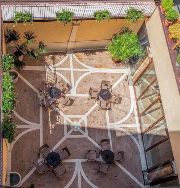 An outdoor patio with a geometric tile pattern, surrounded by potted plants and furnished with round tables and chairs.