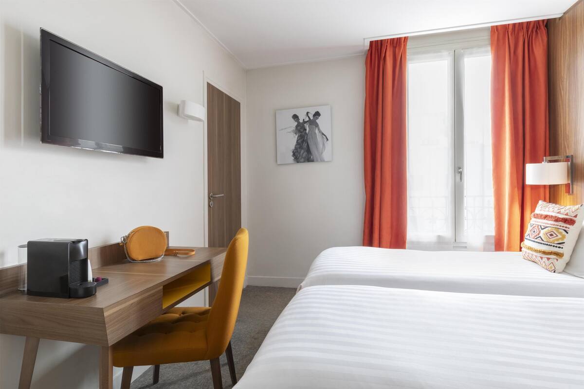 Une chambre d’hôtel équipée d’un téléviseur à écran plat, d’un bureau avec une chaise, d’un lit et d’une fenêtre avec des rideaux.