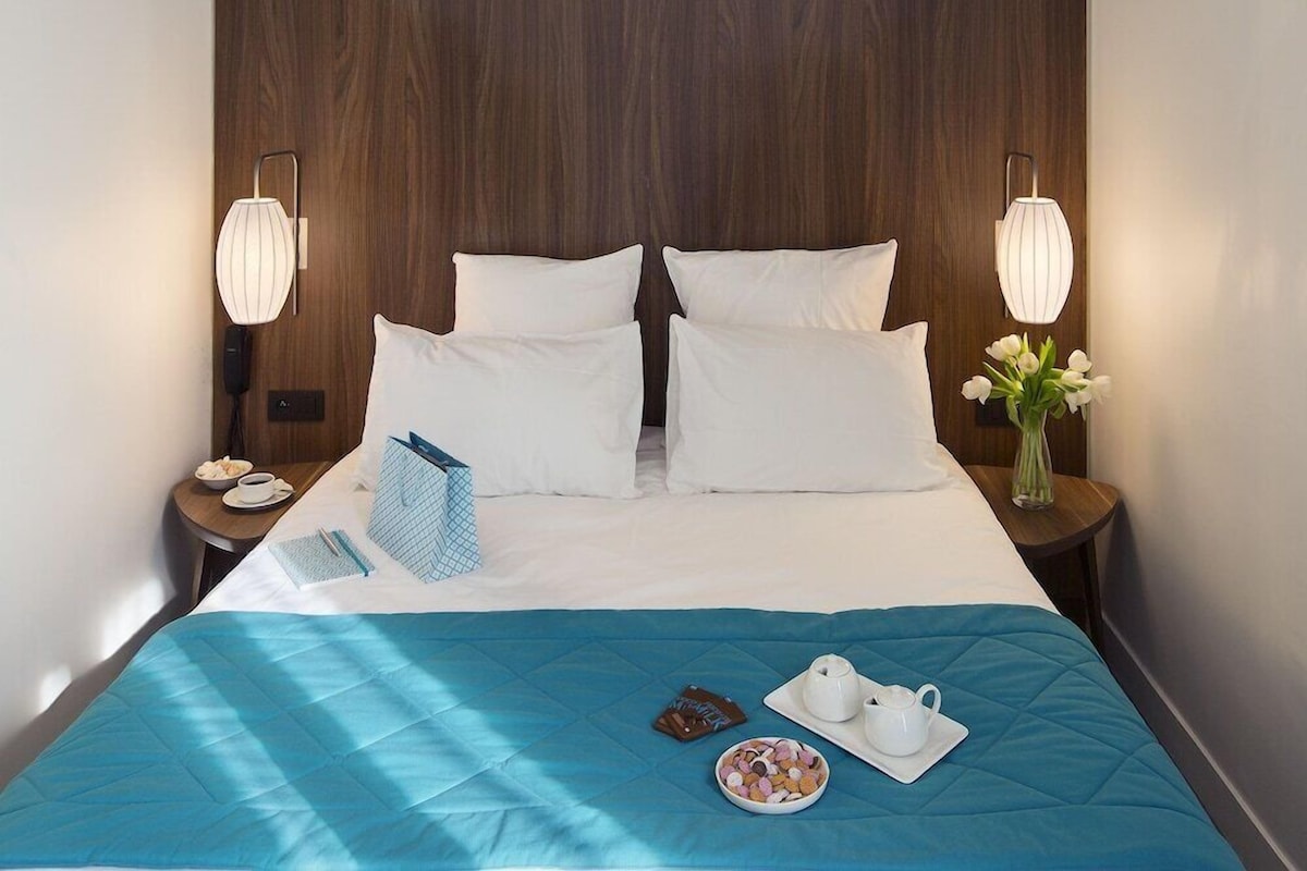 A hotel room with a large bed, two bedside lamps, a bedside table with a phone, a vase with flowers, and a tray with a teapot and cups.