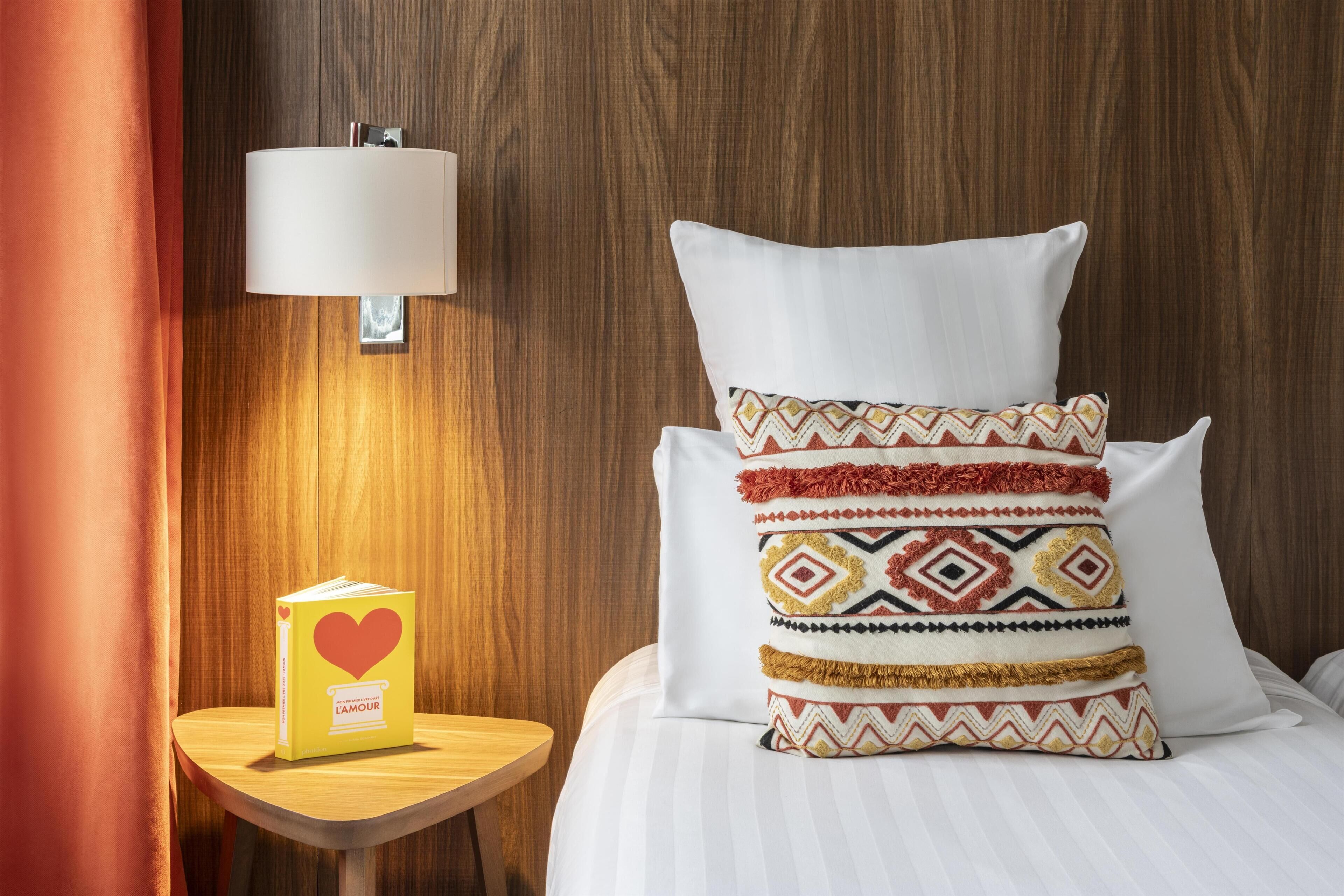A neatly made bed with patterned pillows, a bedside table with a book, and a wall-mounted lamp.
