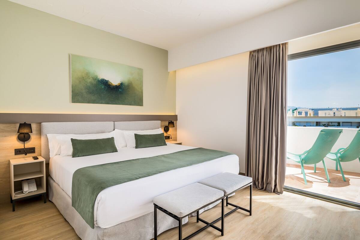 Una habitación de hotel moderna con una cama grande, un banco, una mesita de noche, una lámpara de pared y vistas al balcón.