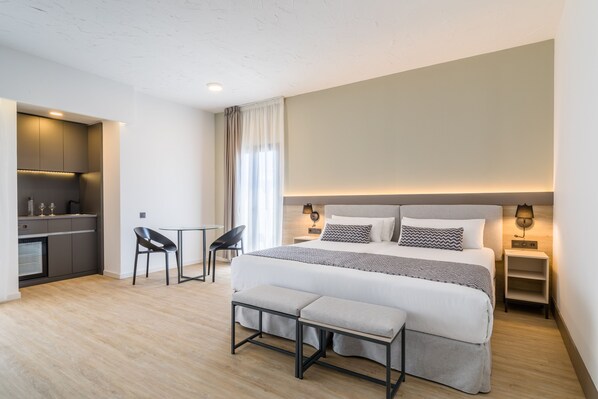 A modern hotel room with a large bed, bedside tables, a desk, and a kitchenette.