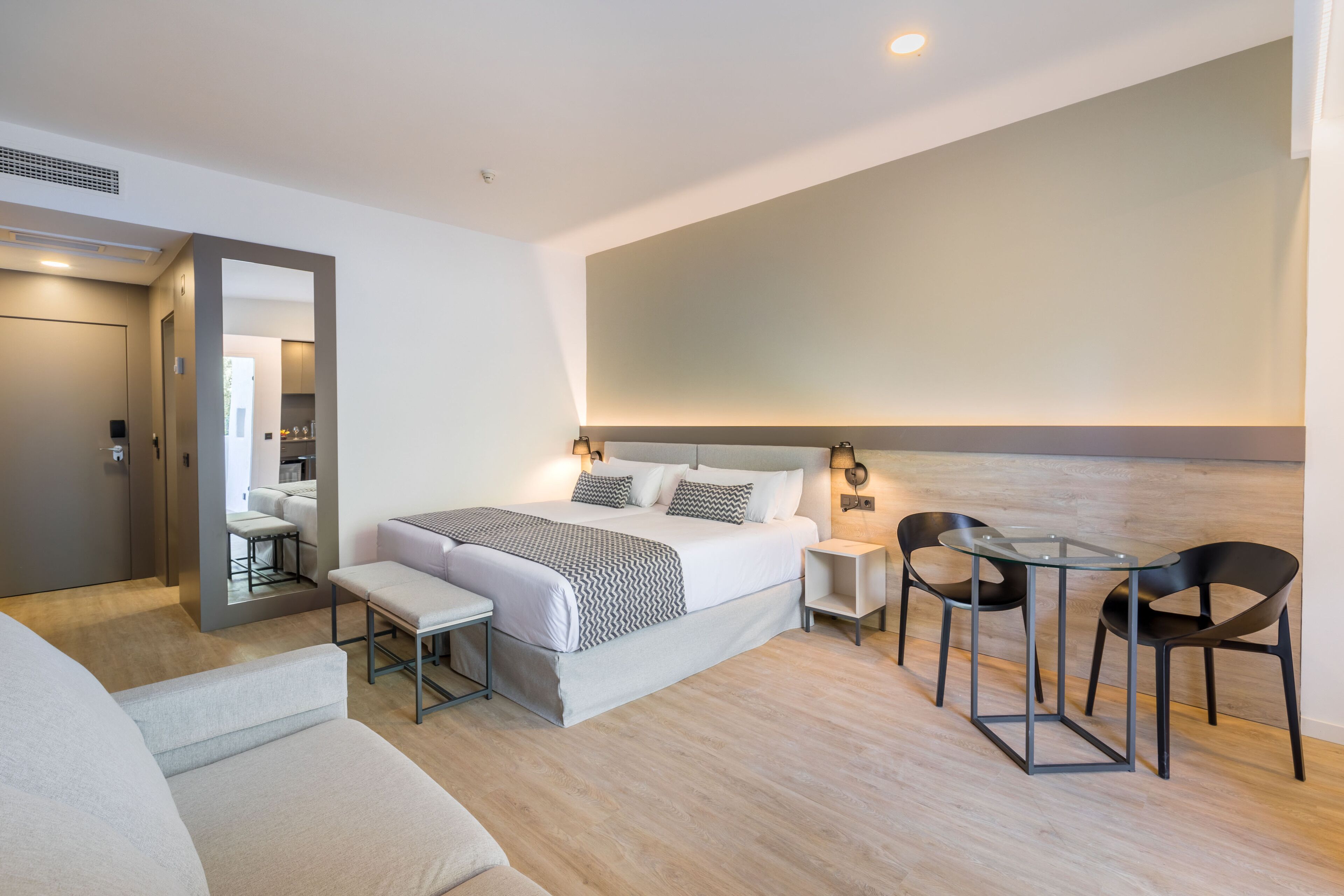A modern hotel room with a large bed, bedside tables, a small table with chairs, a bench, and a mirror.