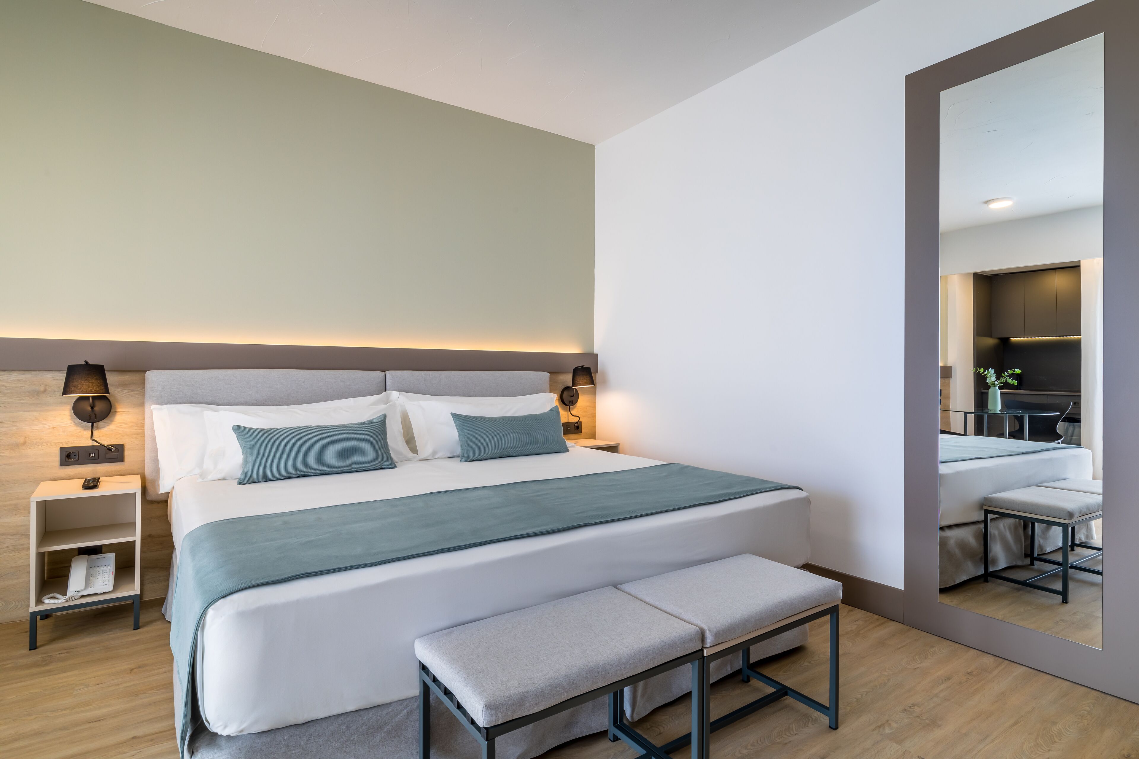 A modern hotel room with a large bed, bedside tables, a bench, and a mirror reflecting the room.