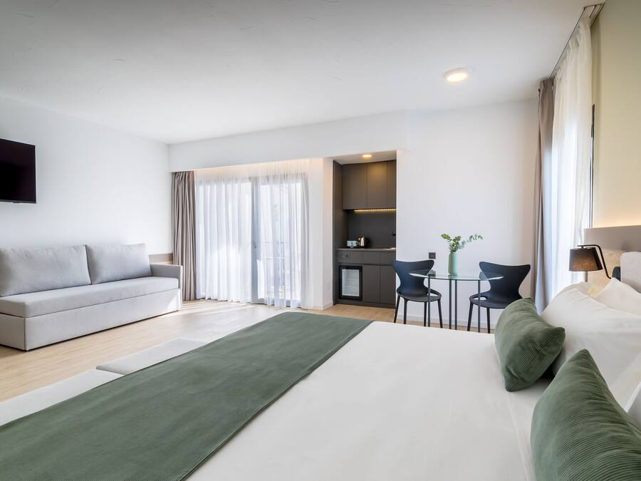 A modern hotel room with a large bed, a sofa, a dining table, and a kitchen area.