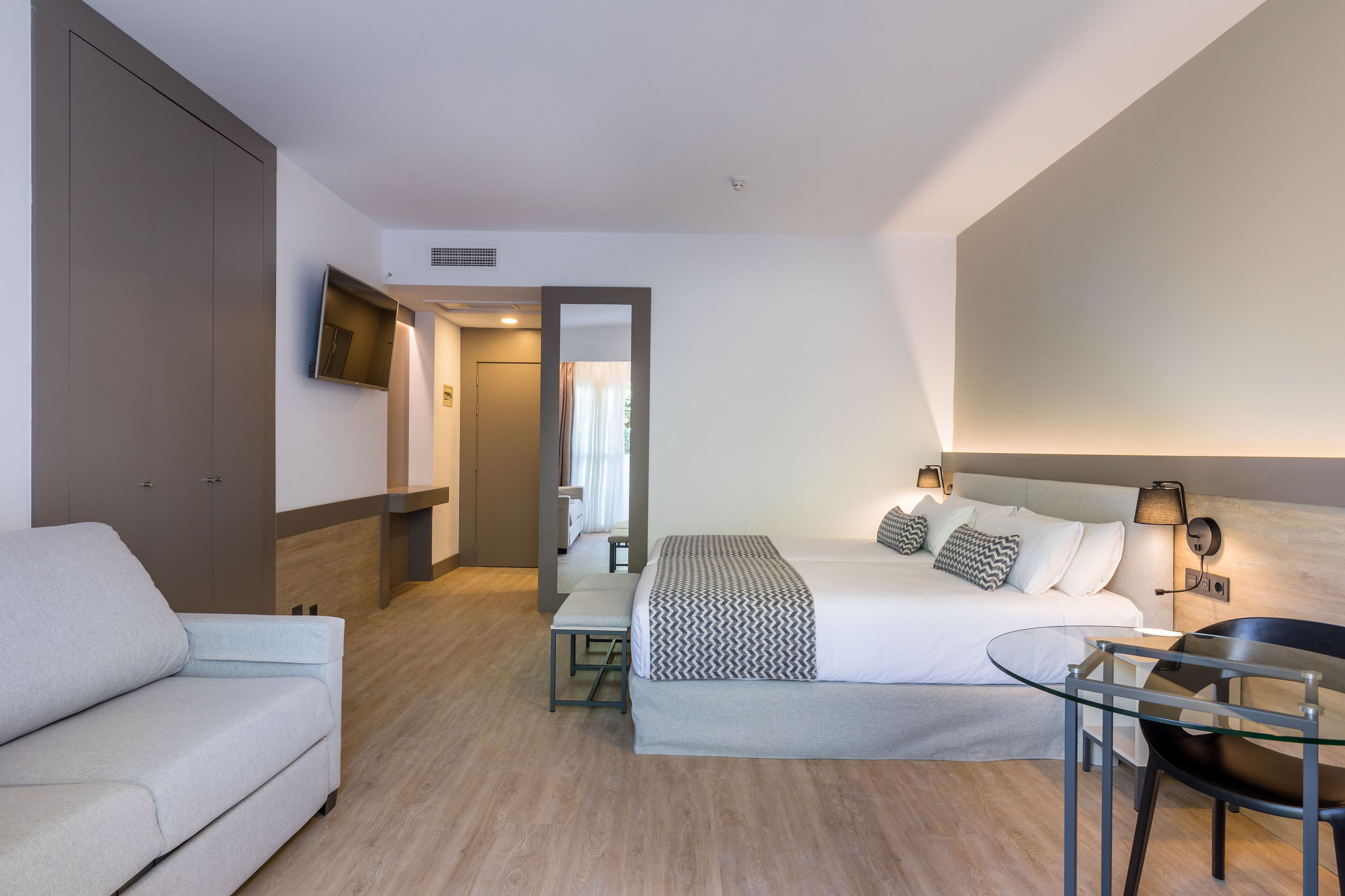 A modern hotel room with a bed, a sofa, a small table, and a TV mounted on the wall.