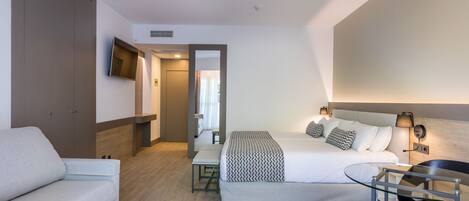 A modern hotel room with a bed, a sofa, a small table, and a TV mounted on the wall.