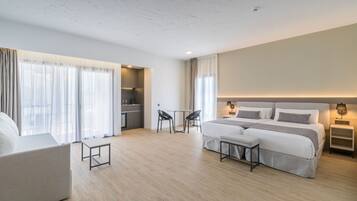 Een moderne hotelkamer met een groot bed, een bank, een klein tafeltje en een bureau. In de hoek bevindt zich een kitchenette en er is uitzicht op de stad door grote ramen.