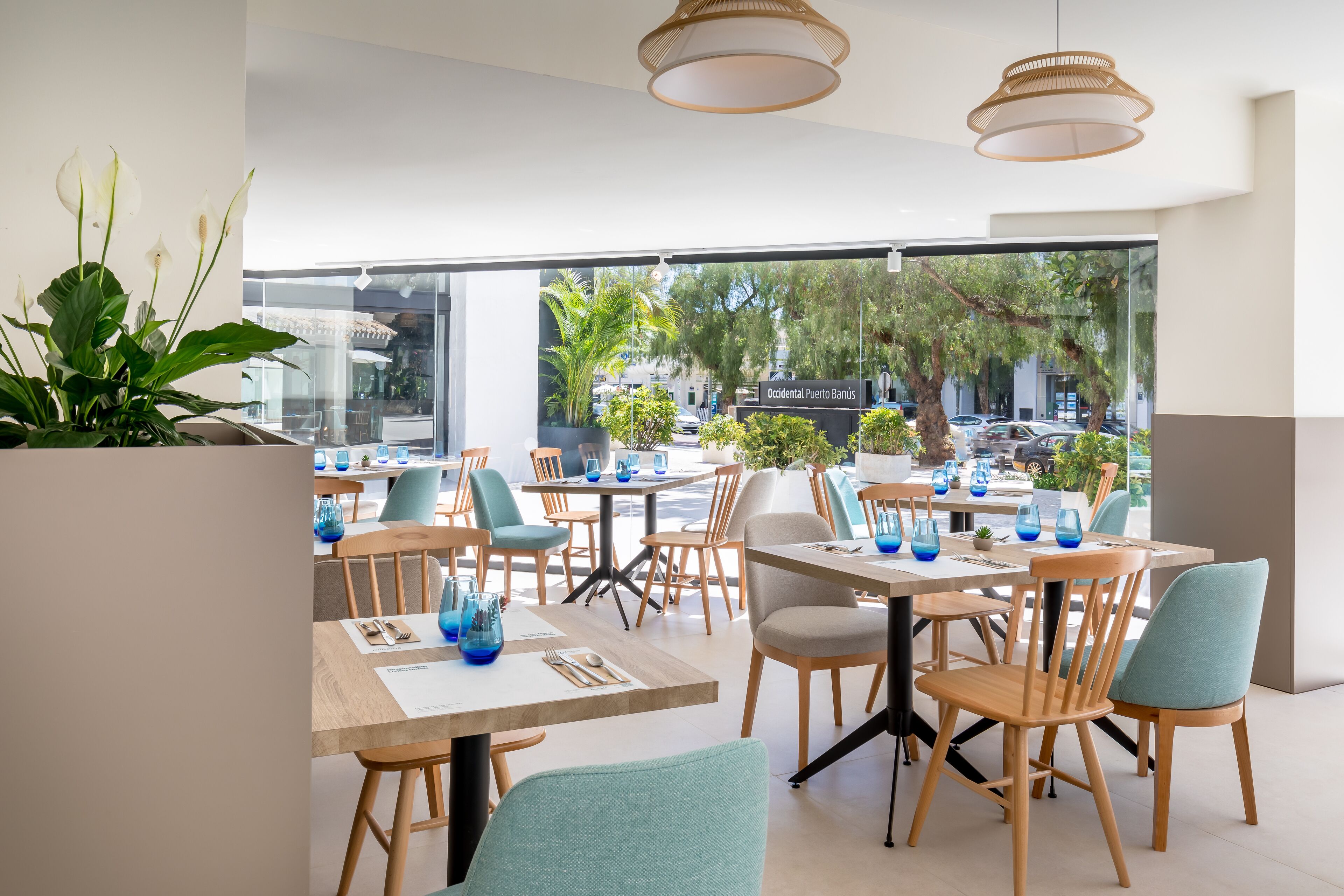 A modern restaurant with wooden tables and chairs, blue glass vases, and large windows offering a view of the street and trees.
