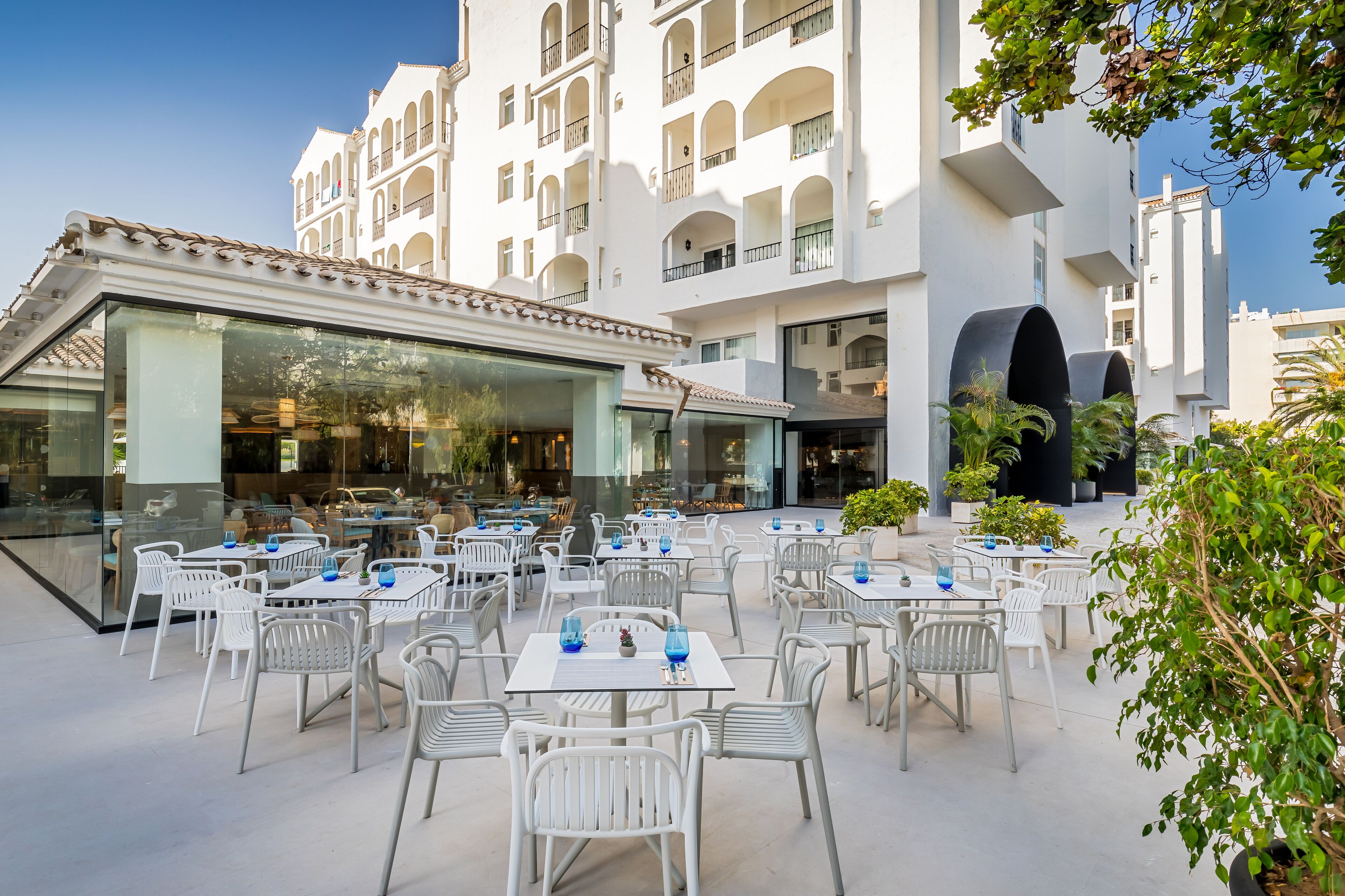 An outdoor dining area with white tables and chairs, surrounded by a white building with balconies and large windows.