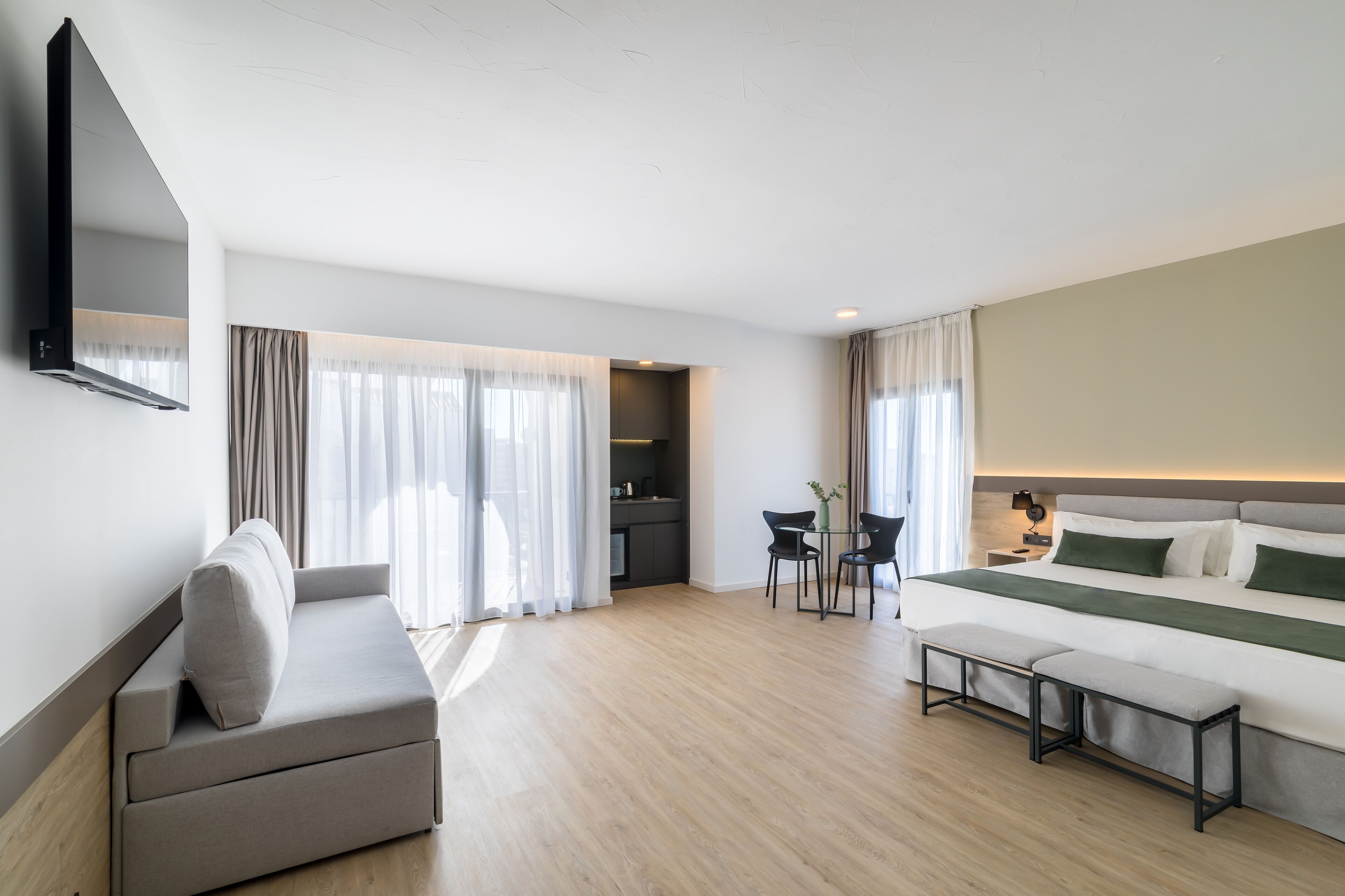 A modern hotel room with a sofa, a bed, a small table, and a TV mounted on the wall.