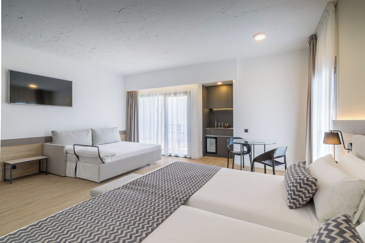 Una habitación de hotel moderna con sofá cama, escritorio, silla y zona de cocina.