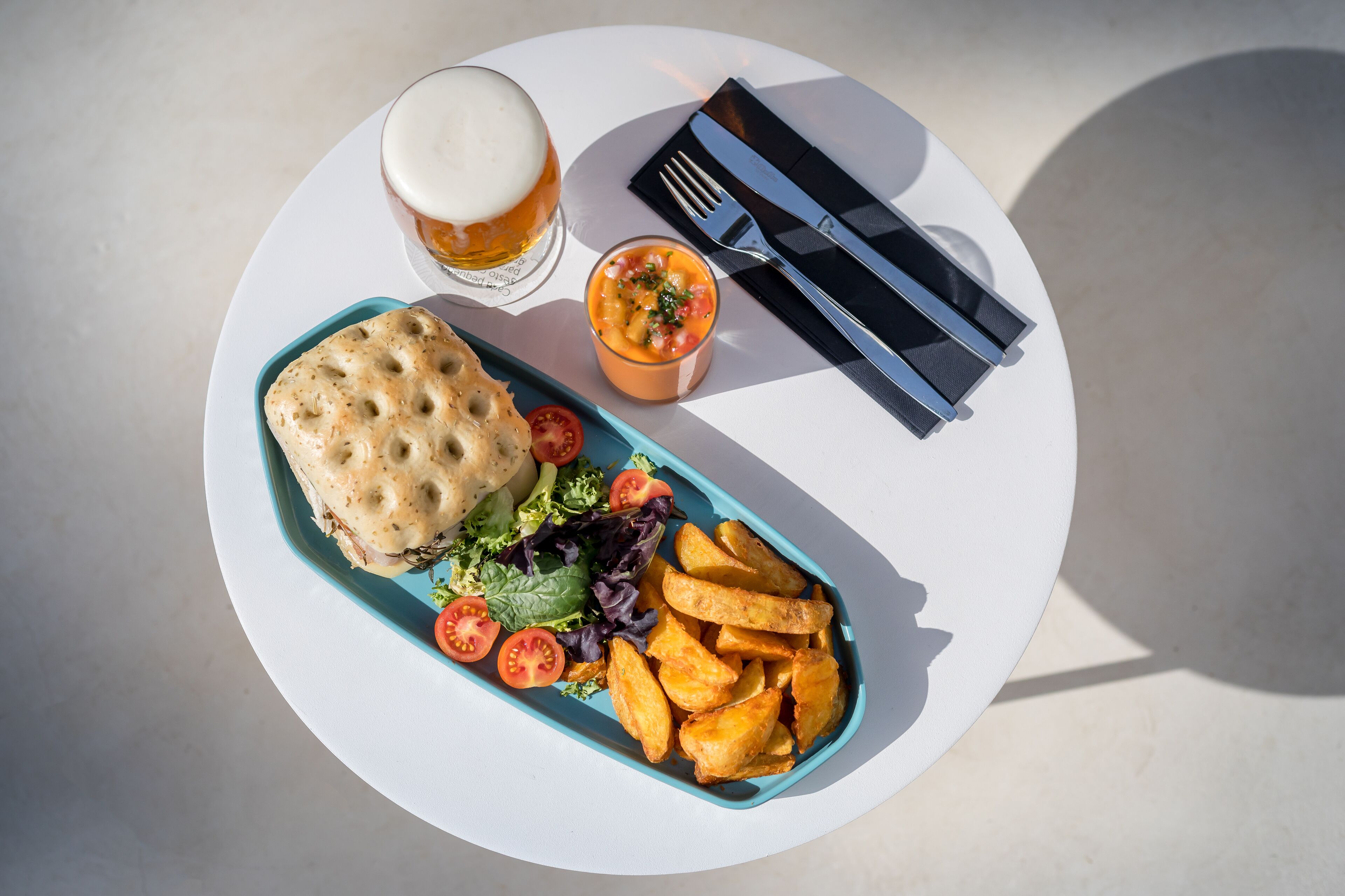A meal consisting of a sandwich, fries, salad, and a small bowl of sauce, served on a white plate with a fork and knife set on a napkin.
