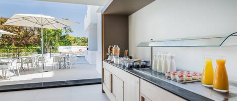 Una barra de desayuno con diversas opciones de comida y bebidas, mesas al aire libre con sombrillas y vistas al verdor.