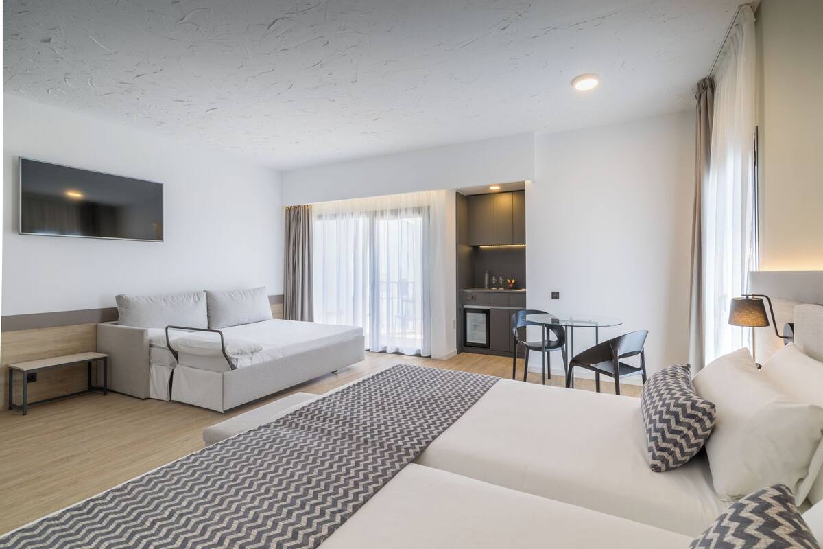 A modern hotel room with a sofa bed, a desk, a chair, and a kitchen area.