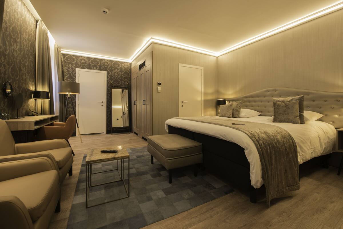 A modern bedroom with a large bed, a sitting area with a sofa and armchair, a small table, and a wardrobe.