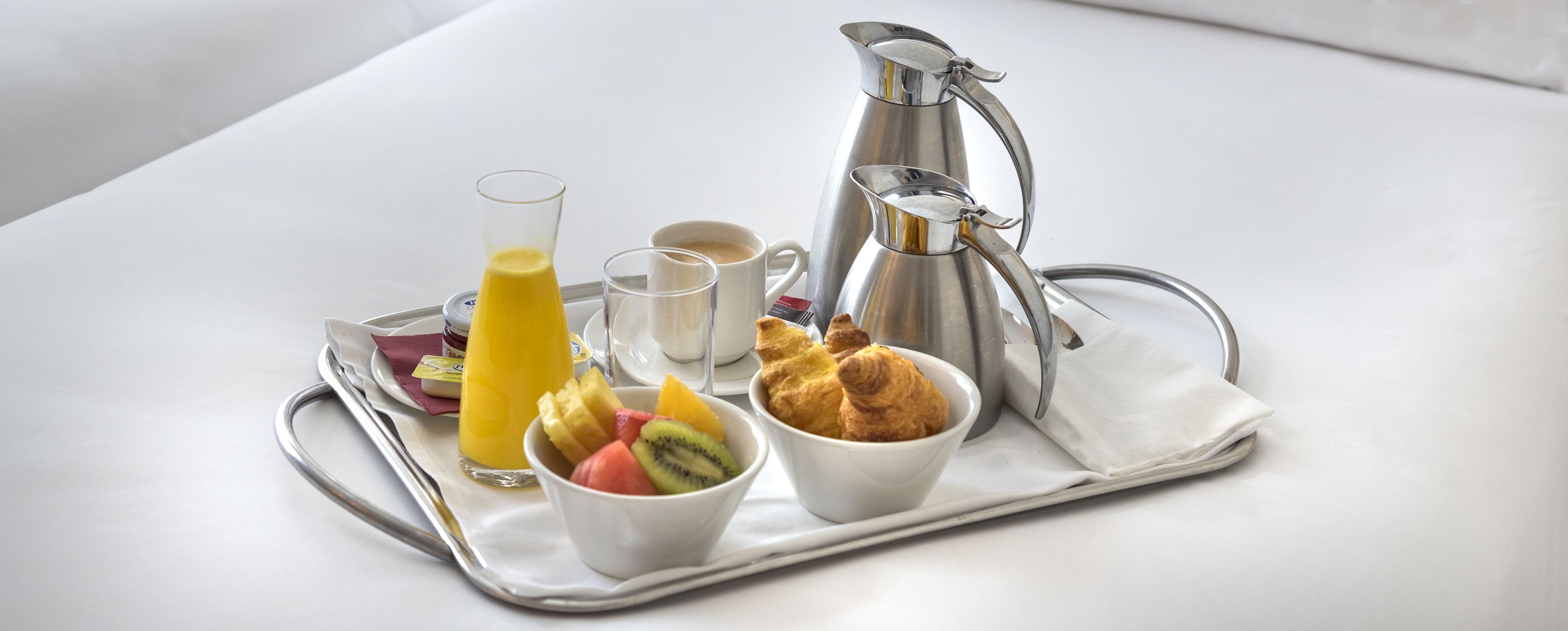 A breakfast tray with croissants, a fruit bowl, a glass of orange juice, a coffee cup, and a silver pitcher.