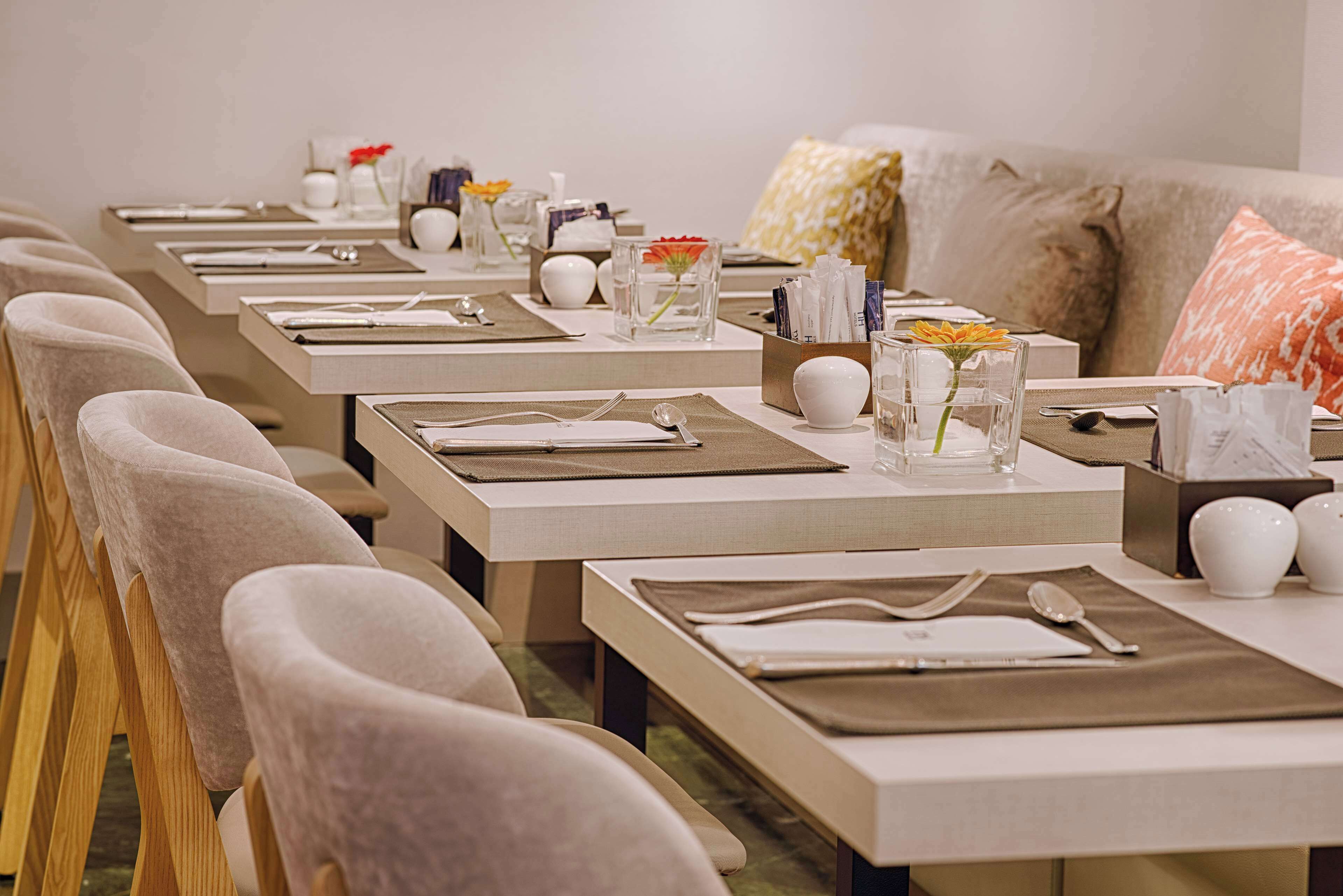 Un comedor con varias mesas dispuestas para el servicio, cada una con un mantel individual, cubiertos y un florero con flores.