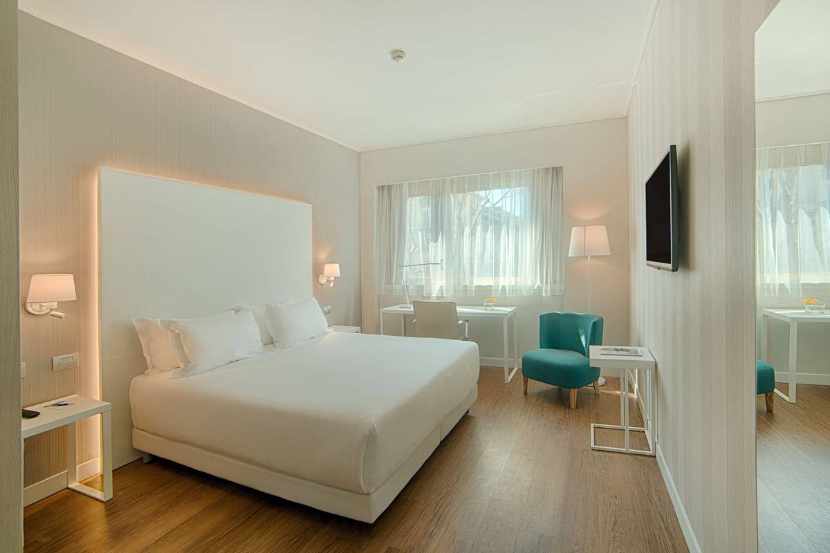 A modern hotel room with a large bed, a desk, a chair, a TV, and a small table.