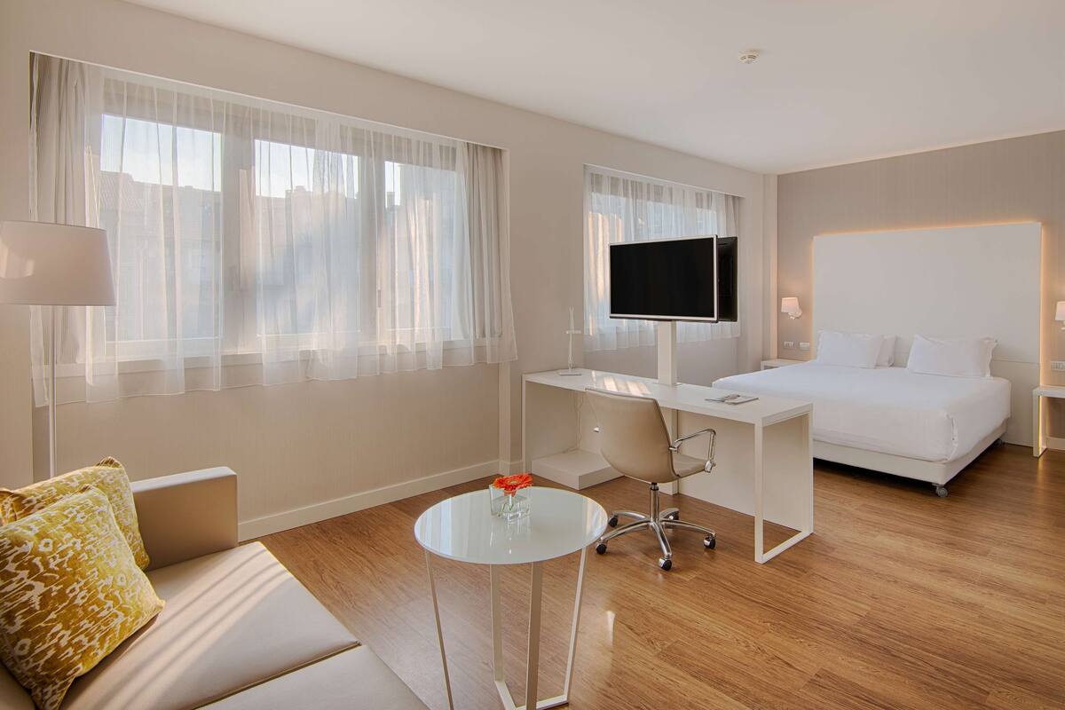 A modern hotel room with a bed, a desk with a flat-screen TV, a sofa, and a small round table with a vase.