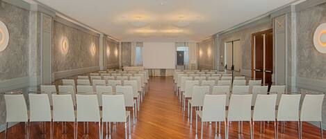 Un salón de conferencias con filas de sillas blancas, piso de madera y una pantalla grande al frente.