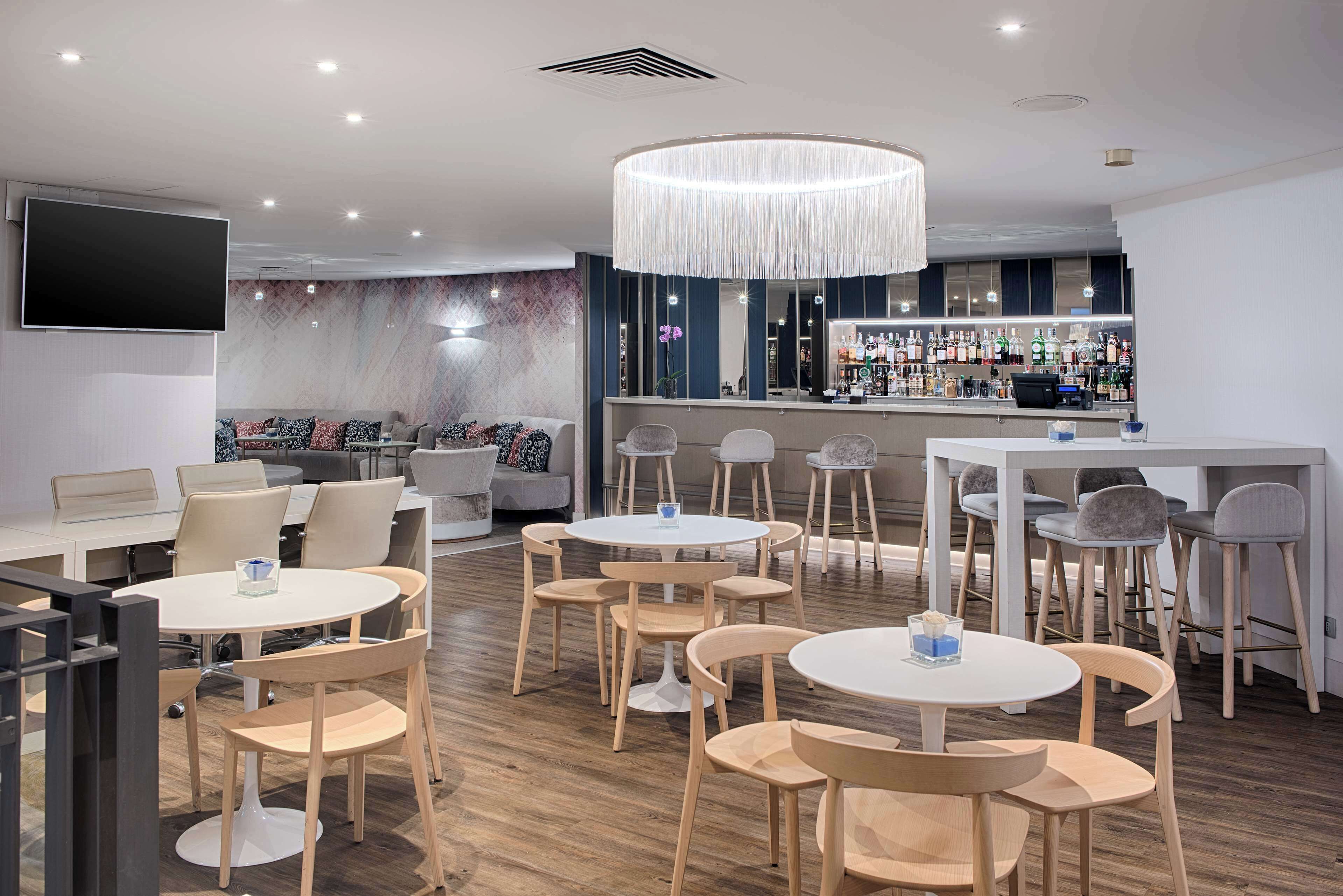 A modern bar area with round tables and chairs, a large TV mounted on the wall, and a bar counter with various bottles.
