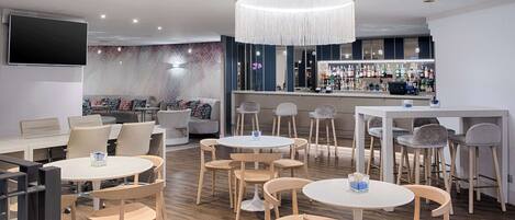 Een moderne bar met ronde tafels en stoelen, een grote televisie aan de muur en een bar met verschillende flessen.