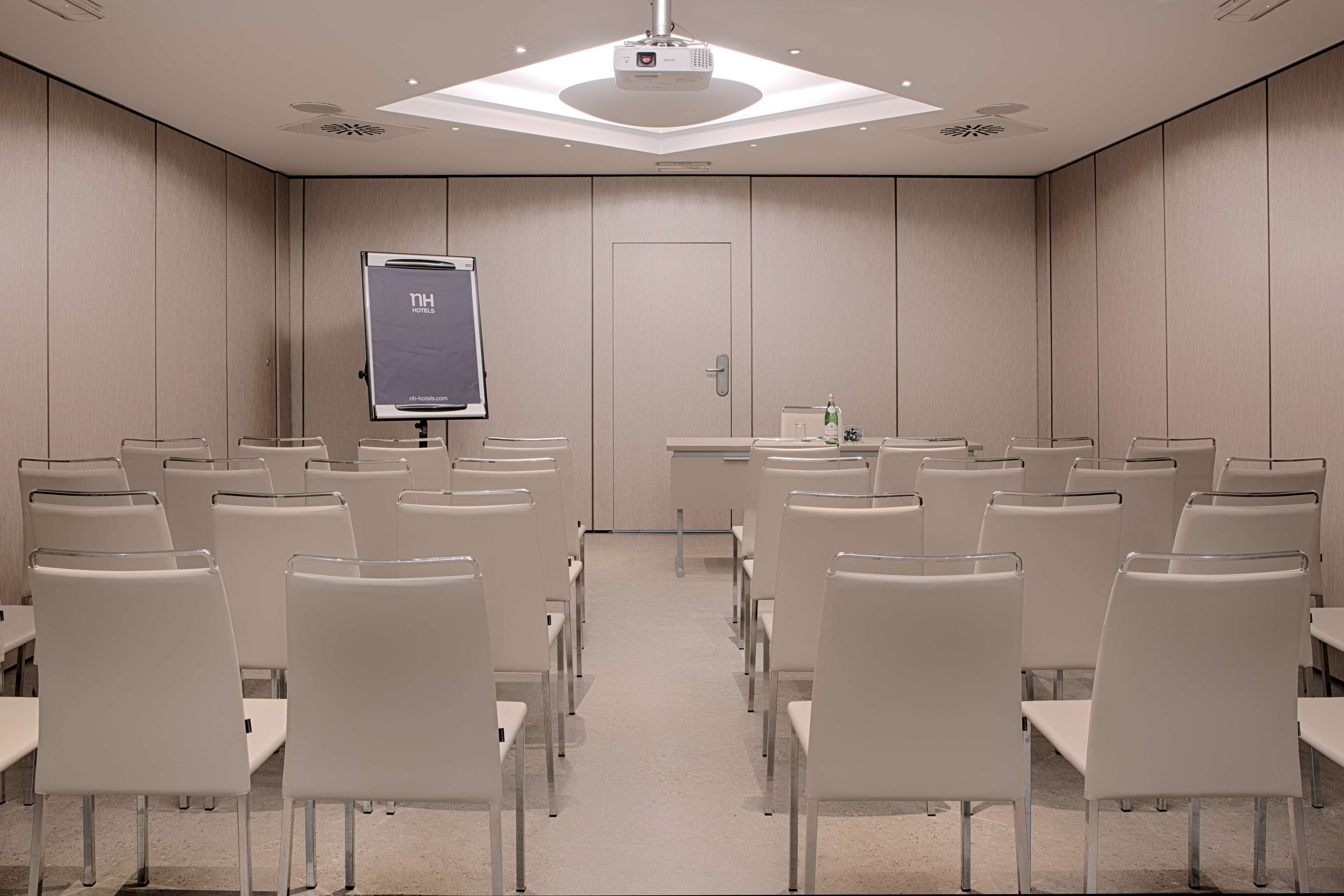 A conference room with rows of white chairs, a podium with a screen, and a door in the center.
