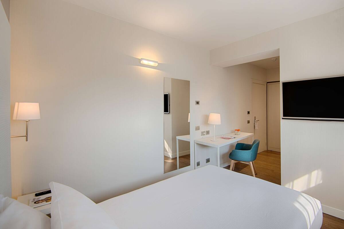 A modern hotel room with a large bed, a desk, a chair, and a flat-screen TV.