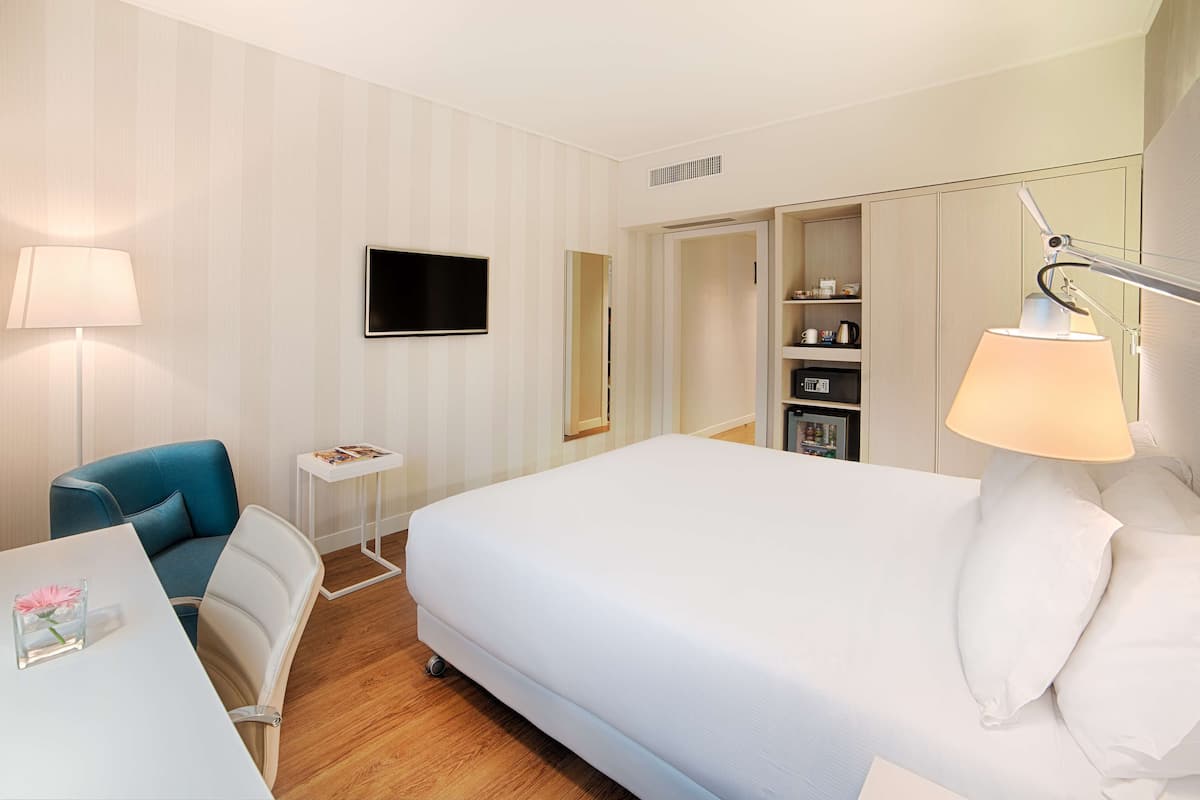 A hotel room with a bed, a desk with a lamp, a chair, a TV, and a minibar.