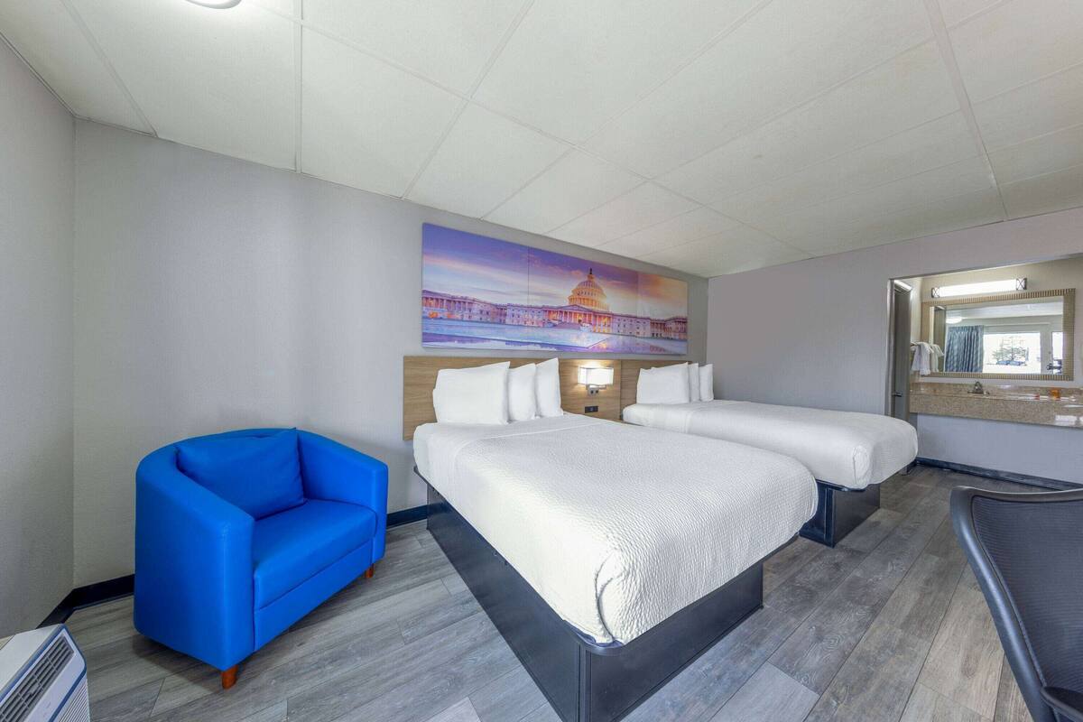 Ein Hotelzimmer mit zwei Betten, einem blauen Sessel, einem großen Gemälde eines Gebäudes und einem Spiegel über dem Bett.