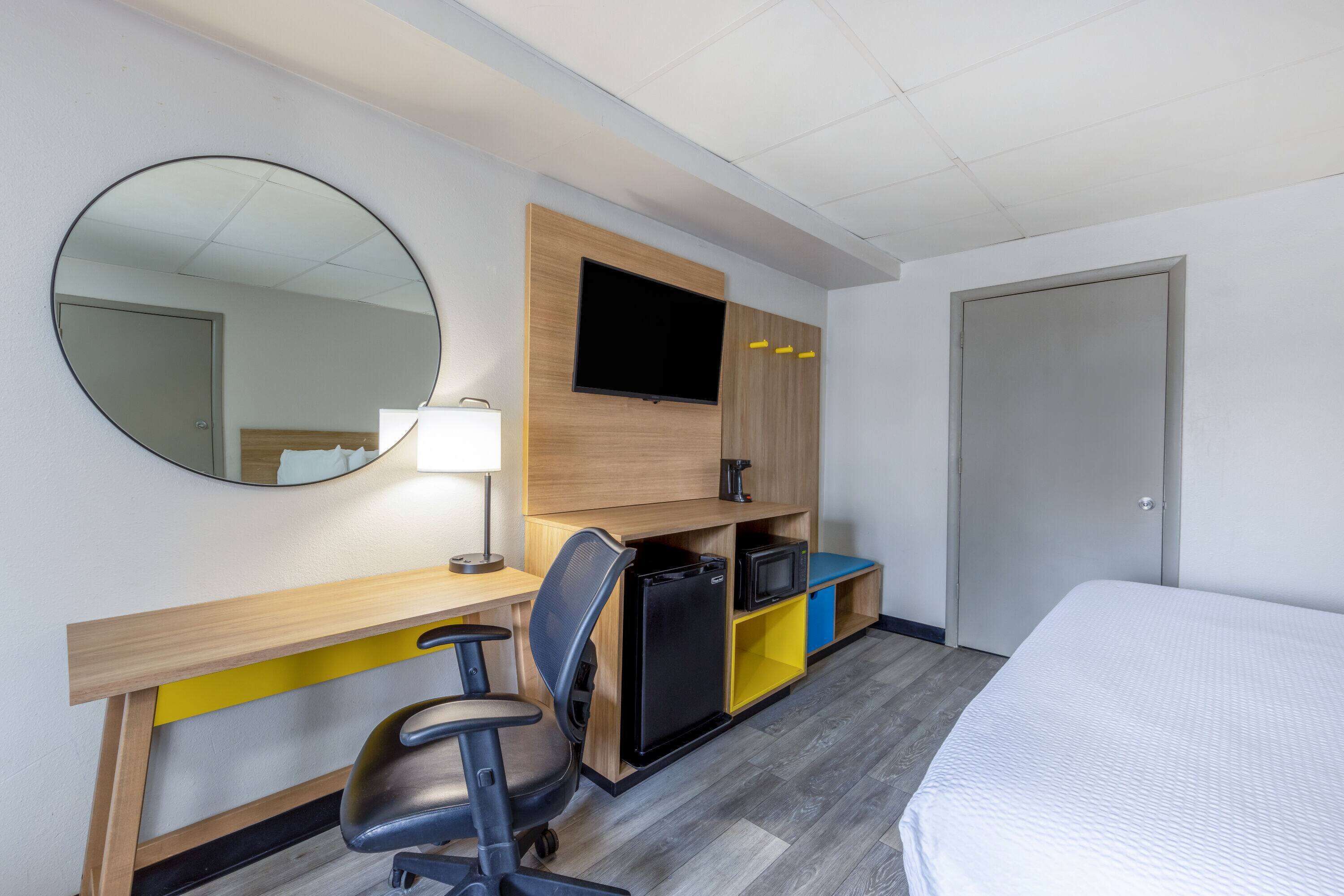 Habitación de hotel con cama, escritorio con silla, refrigerador pequeño, televisor montado en la pared y un espejo.