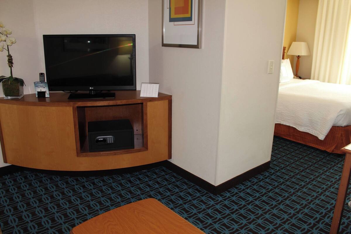 Habitación de hotel con televisión, una caja de seguridad pequeña, control remoto, un cuadro enmarcado y una cama con edredón blanco.