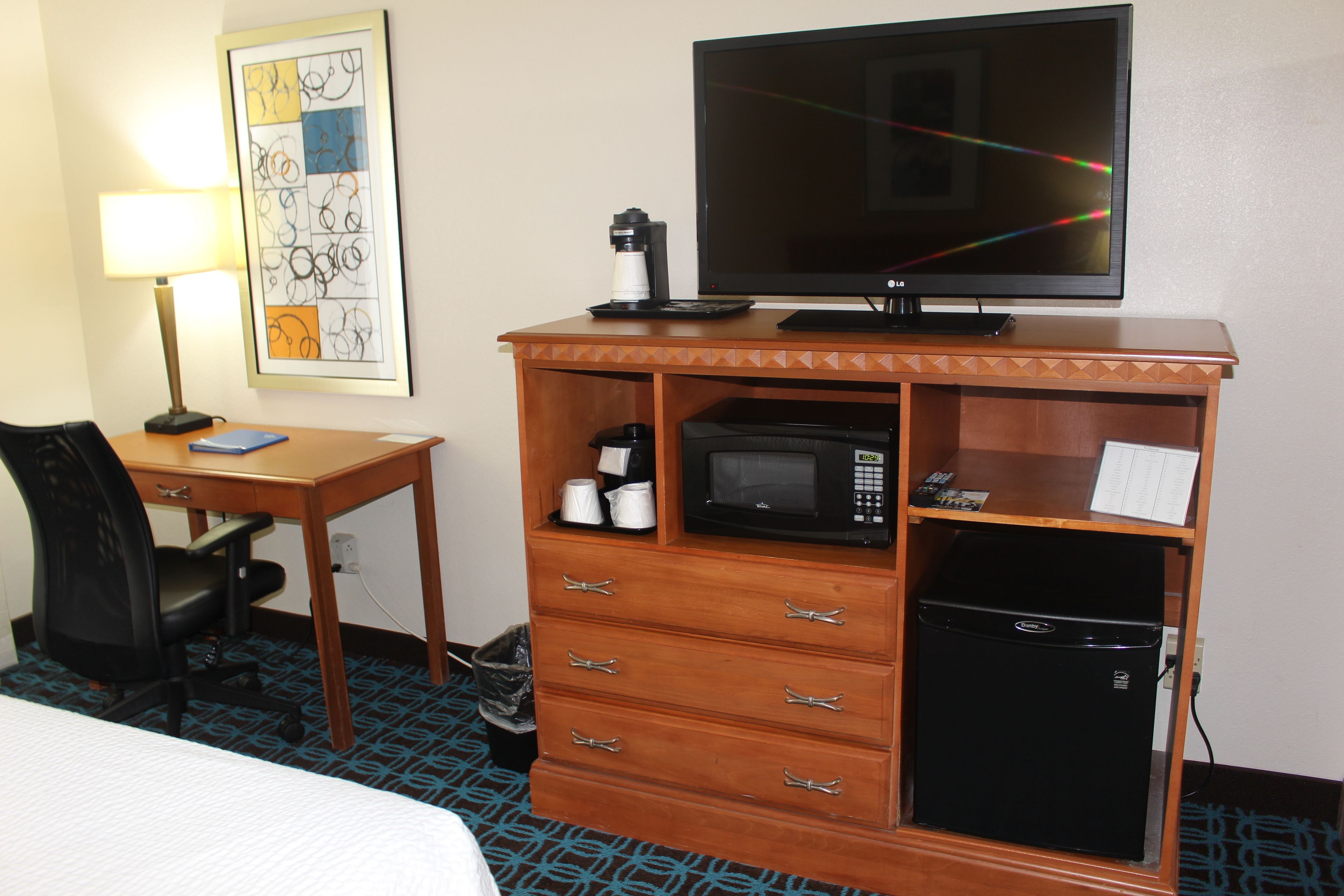 Een hotelkamer met een bed, een bureau met lamp, een televisie, een koffiezetapparaat, een magnetron en een koelkast.
