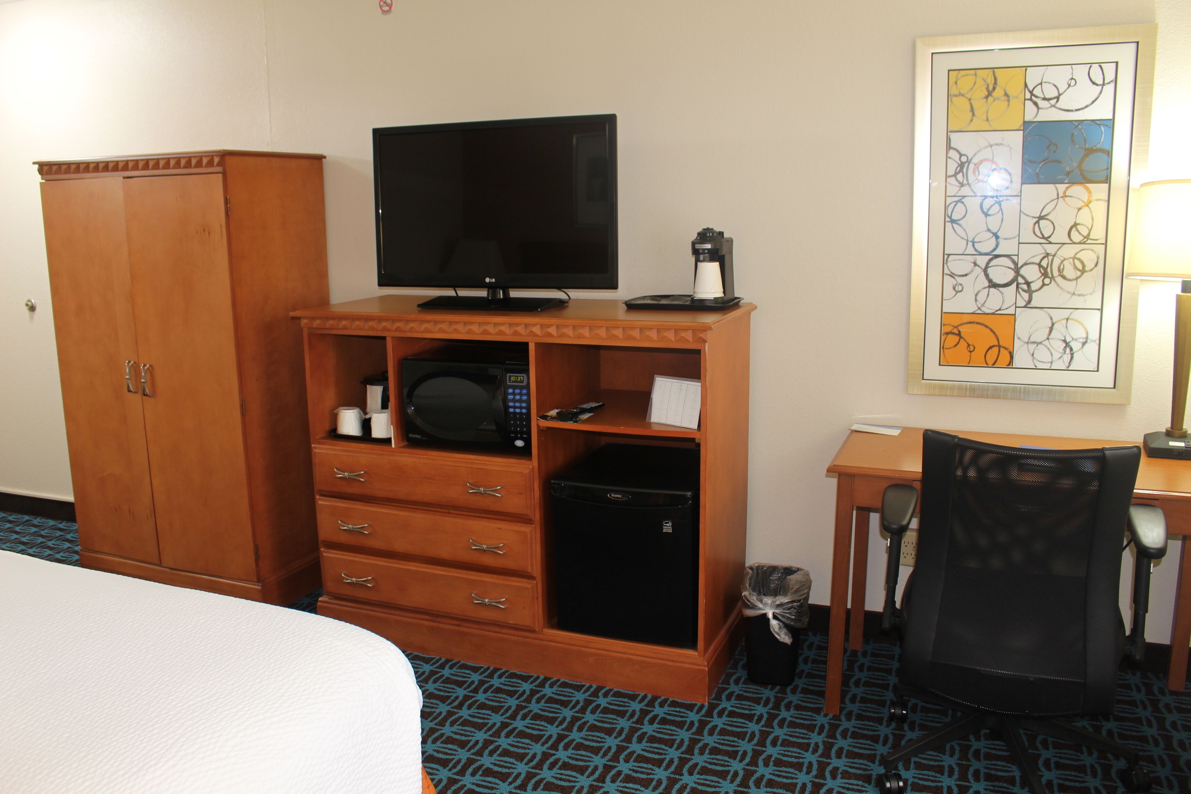 Ein Hotelzimmer mit einem Bett, einem Holzschrank, einem Fernseher, einer Mikrowelle, einem kleinen Kühlschrank, einem Schreibtisch mit Stuhl und einem Wandbild.