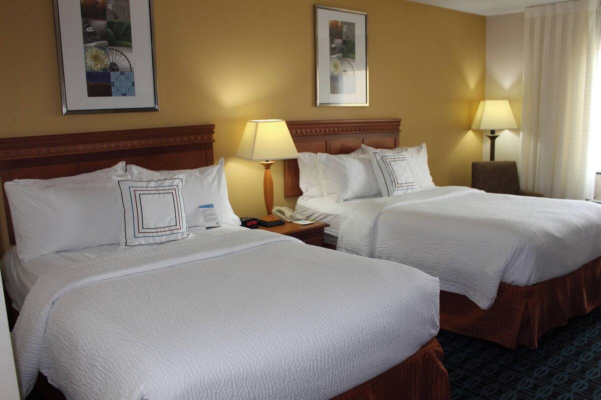 Habitación de hotel con dos camas, cada una con ropa de cama blanca y cabeceros de madera. Hay dos mesitas de noche con lámparas, un cuadro enmarcado en la pared y una ventana con cortinas.